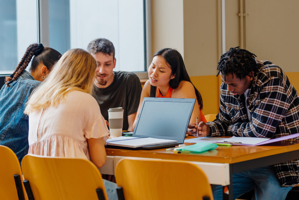 Diverse students in the classroom are collaborating on a project using a laptop and notes.