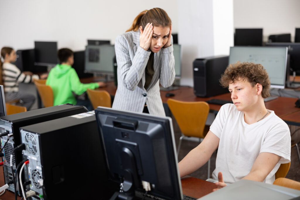 A stressed teacher, gazing at her students' computer screens, feels frustration due to the absence of clear technology guidelines. This situation underscores the challenges for educational leaders.