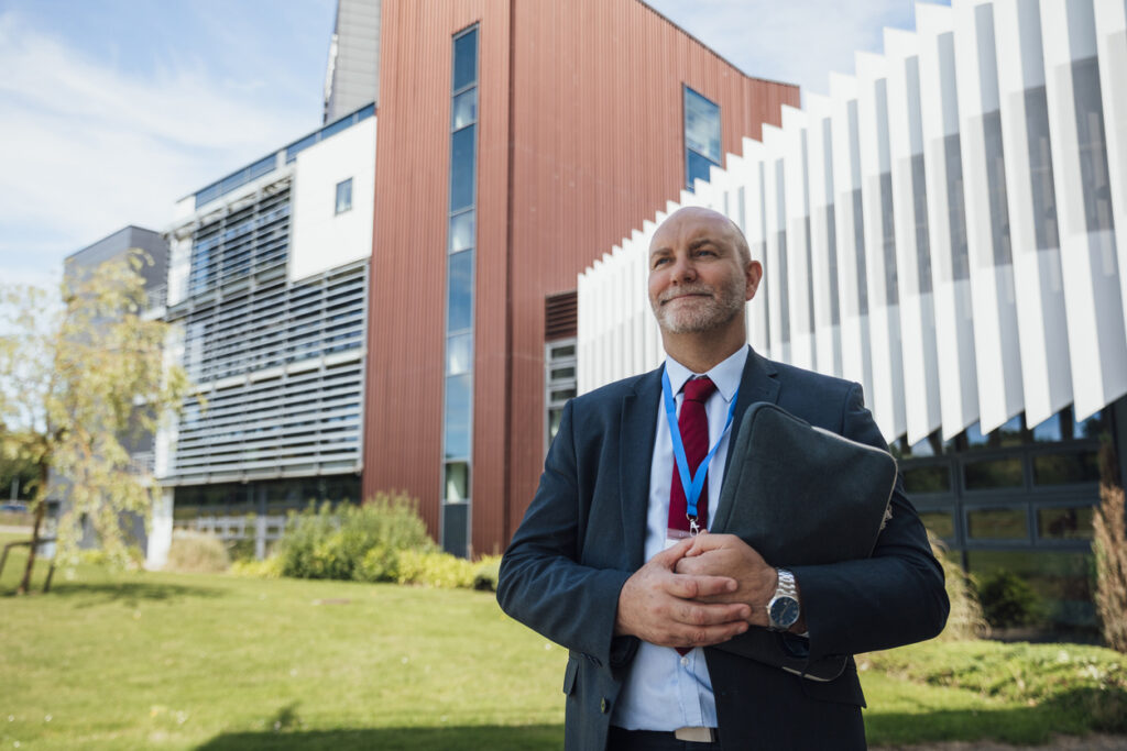 A low-angle view of a proud male school principal holding a file folder standing outside the front of the building